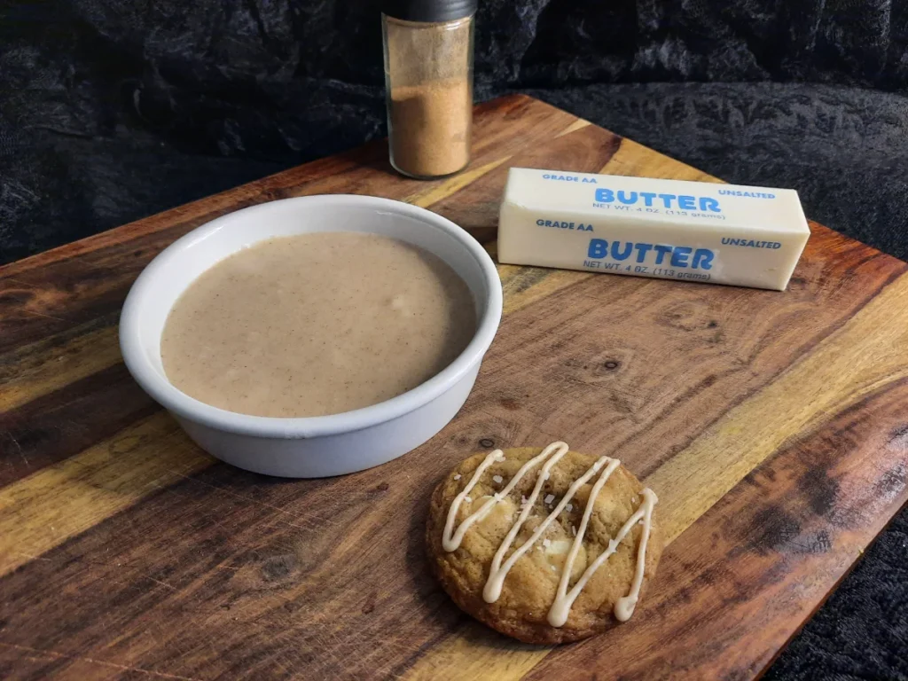 A shallow bowl of spiced icing with a cookie drizzled with icing beside it. There's butter and a chai spice mix in the background.