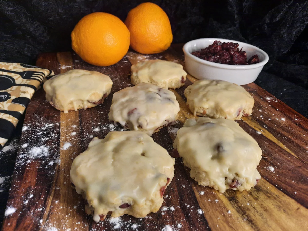 Cranberry Orange Scones Cranberry scones covered in an orange glaze spread out on a lightly floured cutting board. In the background there's a napkin, two oranges and a bowl of dried cranberries.